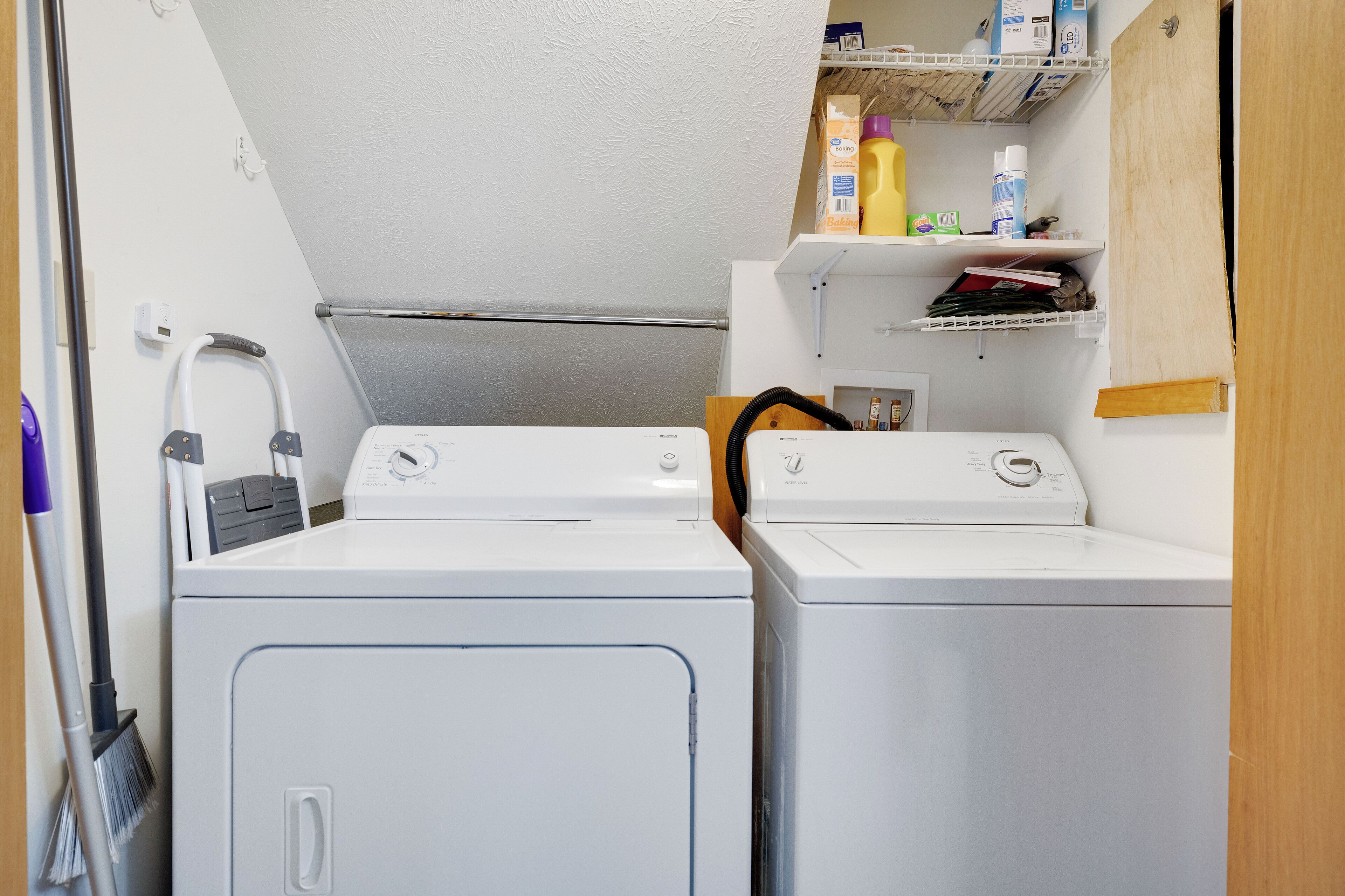 Een wasruimte met twee witte wasmachines, een bezem en planken met schoonmaakspullen.