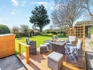Een terras buiten met wicker meubilair, een houten terras en uitzicht op een tuin en bomen.