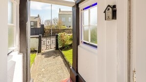 A covered outdoor corridor with a gravel path leading to a gate, a view of a residential area, and a door with a window and a doorbell. - 3 bedroom accommodation in Silecroft, near Millom (Silecroft, near Millom)