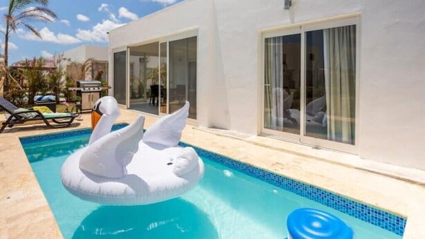 A swimming pool with a swan-shaped inflatable float, a white building with large windows, and a palm tree in the background. - Villa Paradise Punta Cana (Punta Cana)