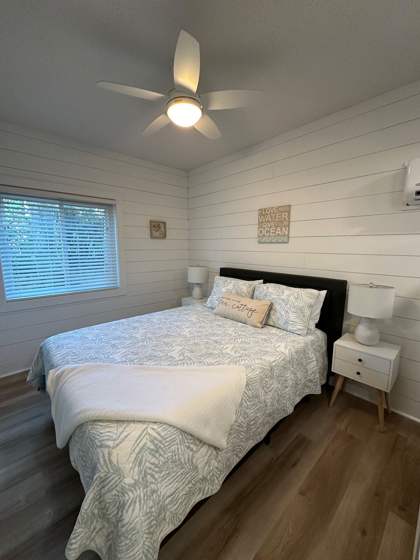 Un dormitorio con cama, ventilador de techo, mesita de noche con lámpara, ventana con persianas y una pared con un letrero decorativo.