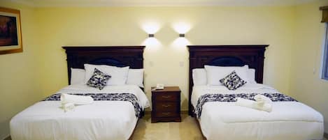 A hotel room with two beds, each with a wooden headboard, white bedding, and decorative pillows. There is a small bedside table between the beds.