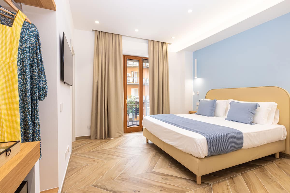 A bedroom with a bed, wooden floor, curtains, a TV, and a view of the outside.