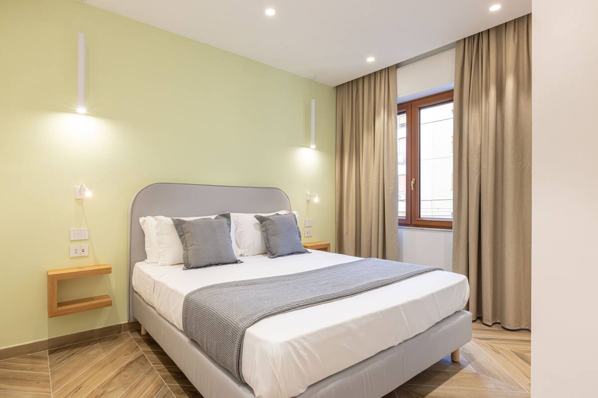 A neatly made bed with a grey headboard, white bedding, and a grey throw blanket. There are two bedside tables with lamps, and a window with beige curtains.