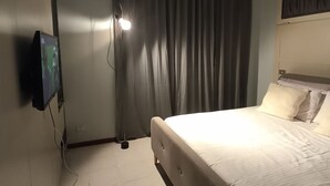 Een hotelkamer met een bed, gordijnen, een aan de muur bevestigde televisie en een plafondlamp.
