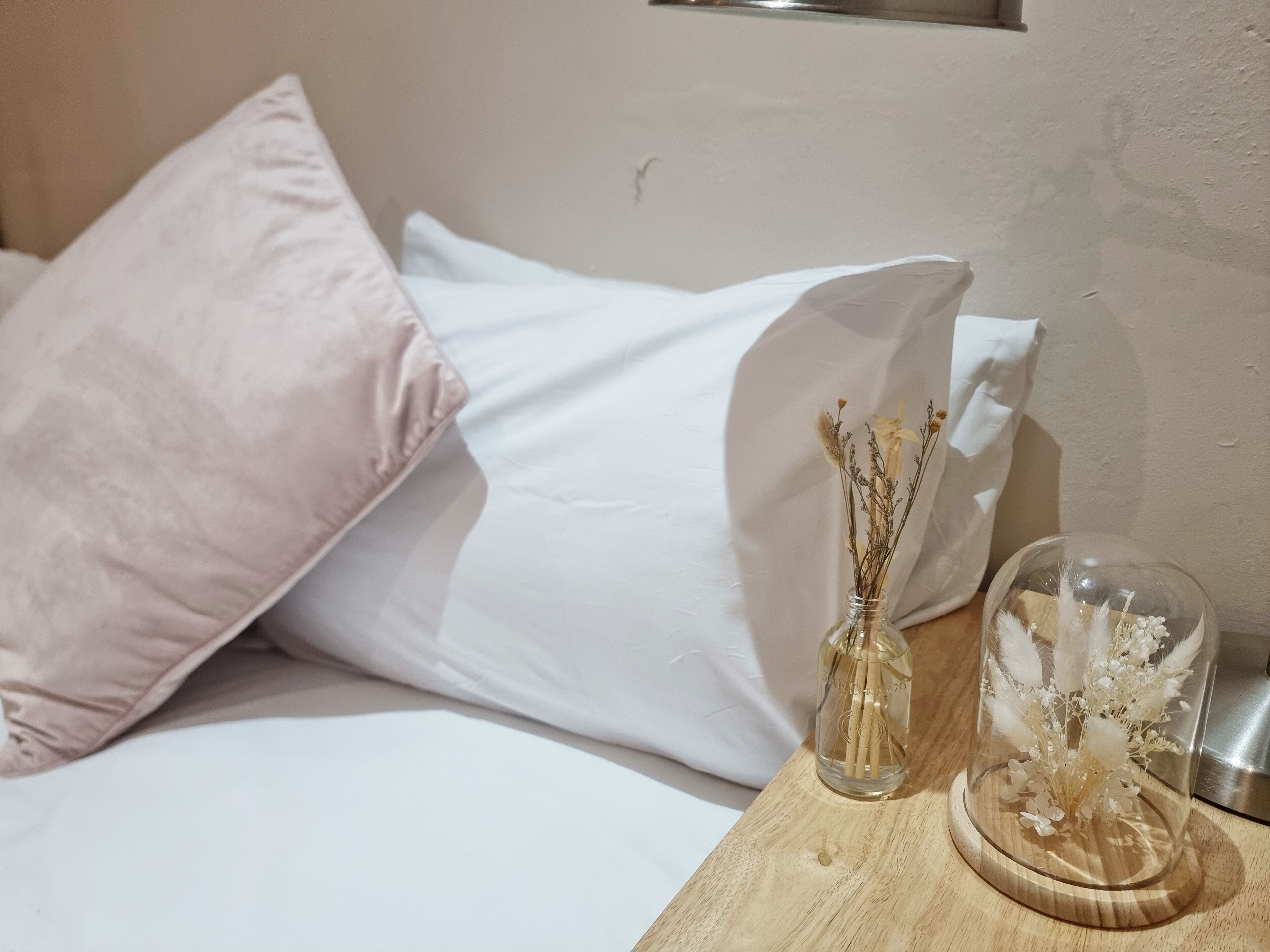 A neatly made bed with white and pink pillows, a clear glass vase with dried flowers, and a wooden nightstand.