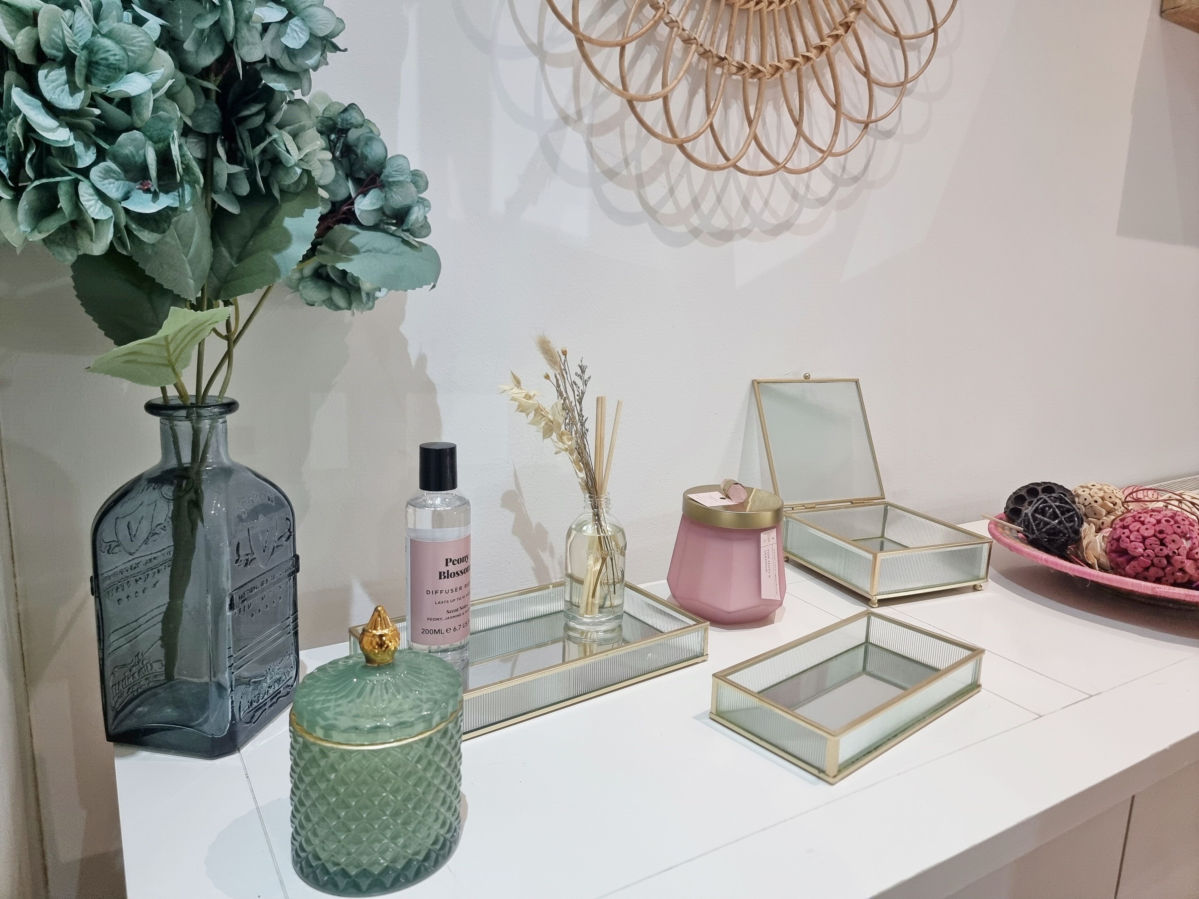 Una encimera blanca con un florero de hojas verdes, una botella de perfume, un recipiente de vidrio con tapa dorada y un candelabro rosa.