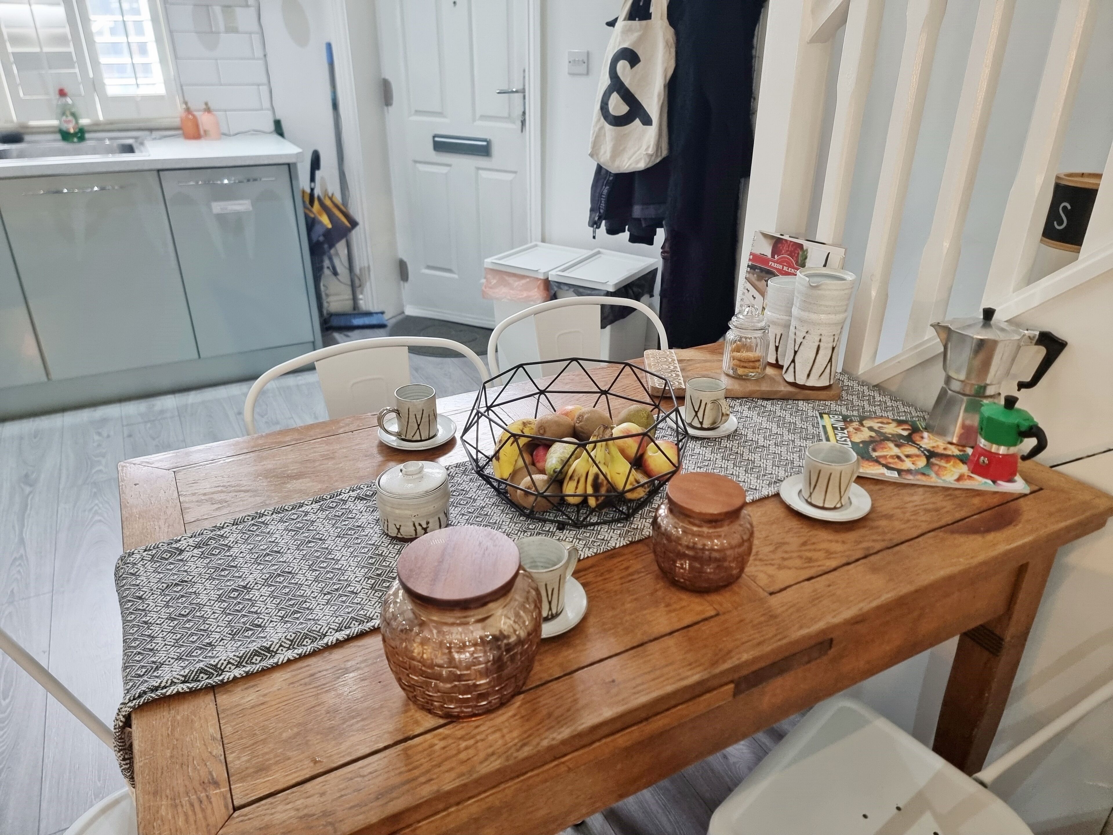 Uma mesa de jantar de madeira com um jogo americano estampado, uma cesta de frutas e diversos utensílios de cerâmica e vidro.