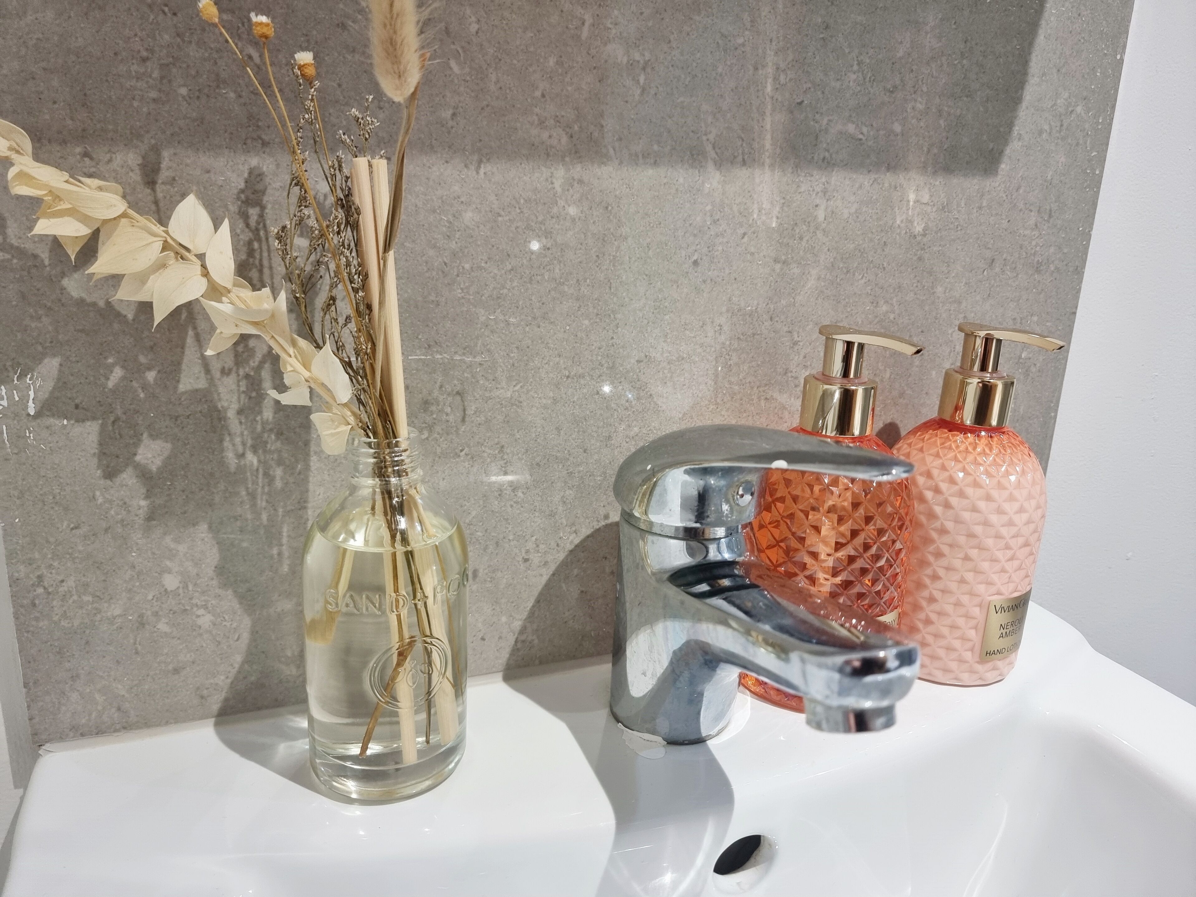 A bathroom sink with a clear glass vase containing dried flowers and a pump bottle of hand lotion.