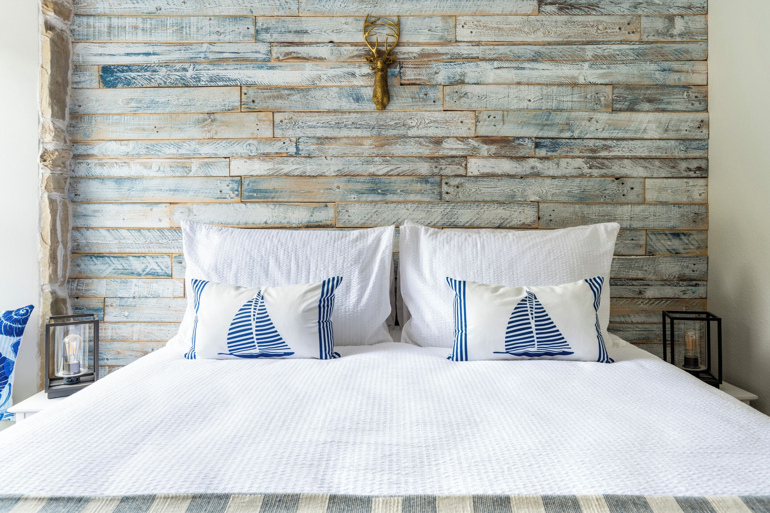 A bed with white bedding and striped pillows, a rustic wooden headboard, and a mounted deer head.