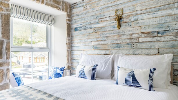 A bedroom with a rustic wooden wall, a large bed with white and blue bedding, and a window with a striped valance. - Beachspoke Loft (Redruth)