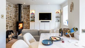 A cozy living room with a stone wall, a fireplace, a flat-screen TV, and a dining area with a table set for two. - Beachspoke Loft (Redruth)