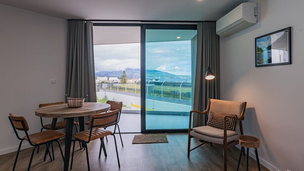 A modern living room with a round wooden table, four chairs, a sofa, and a large sliding glass door leading to a balcony with a city view. - Casa ProMar - Sea View Serenity (Ponta Delgada)