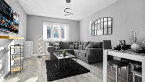 A modern living room with a grey sofa, a glass coffee table, and a dining table with chairs. There is a TV mounted on the wall displaying movie posters. - Hotel Comforts & Home Conveniences -sleeps 8 (Birmingham)