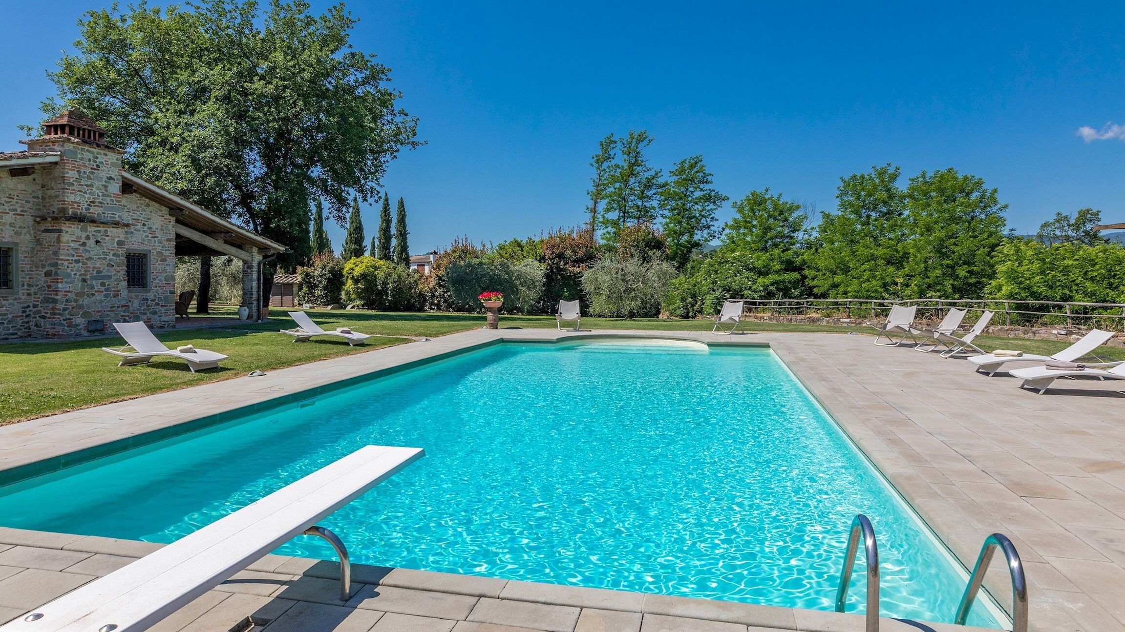 A rectangular swimming pool with a stone building in the background, surrounded by greenery and a clear sky.