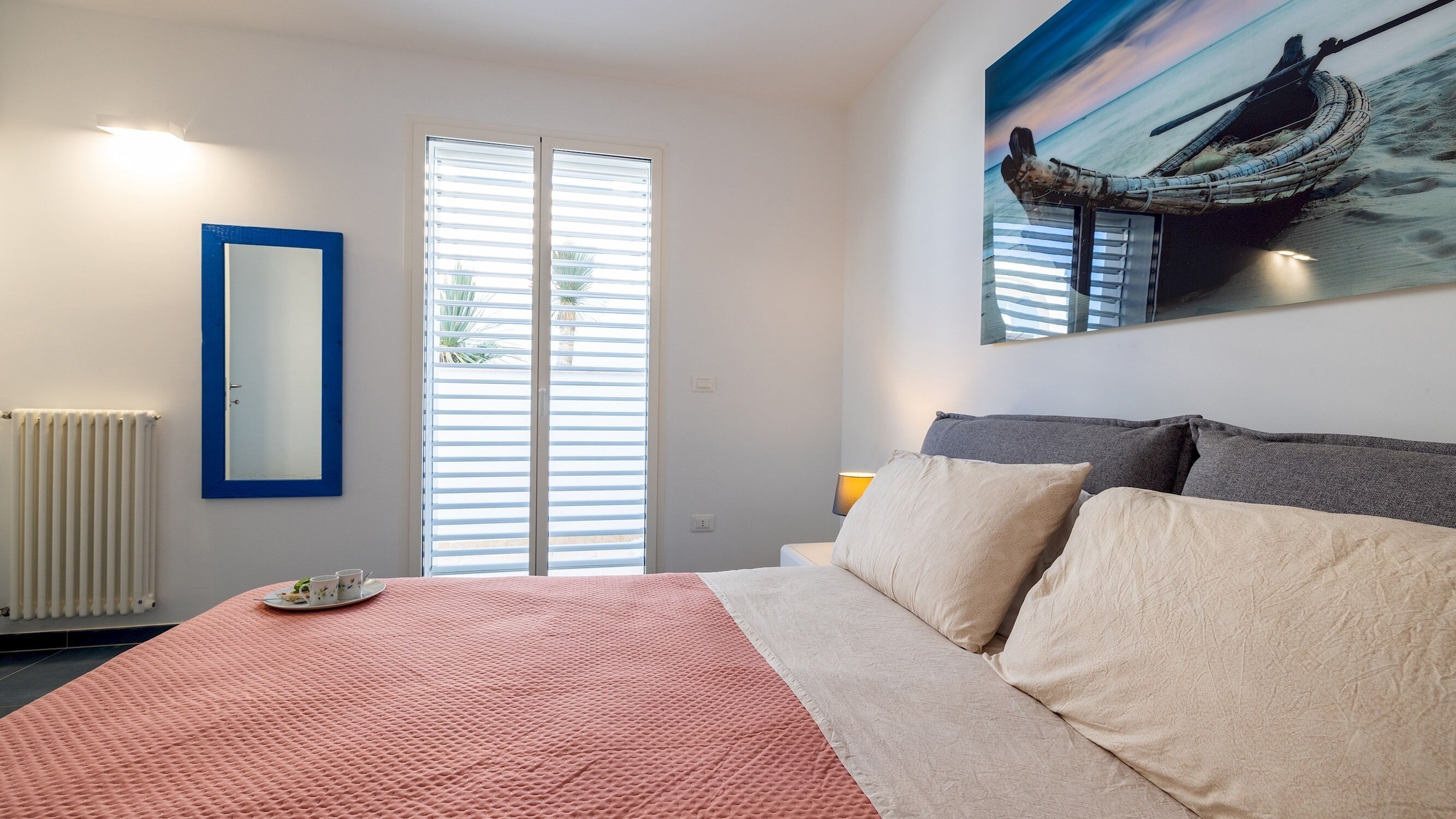 A bedroom with a bed, a radiator, a window with blinds, a painting of a boat, and a bedside table with a lamp.