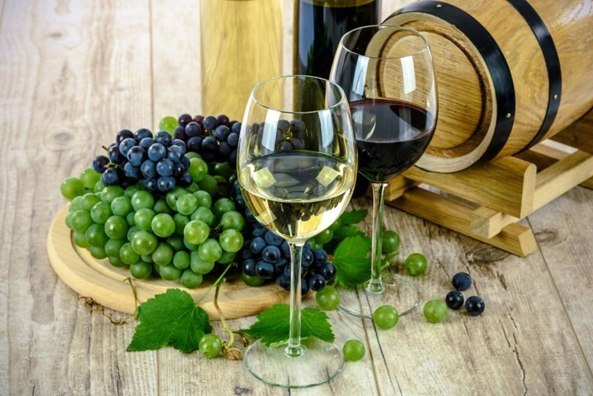 A wooden barrel, two wine glasses, and a wooden board with green and dark grapes.