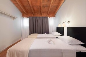 A hotel room with two beds, wooden ceiling beams, a white wall, a window with curtains, and a nightstand with a lamp. - Agrology Lux (Rhodes)