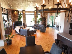 A spacious living room with a stone wall, wooden flooring, and a fireplace. There are multiple seating areas, including a sofa and armchairs, and a dining table with stools. - Agrology Lux (Rhodes)