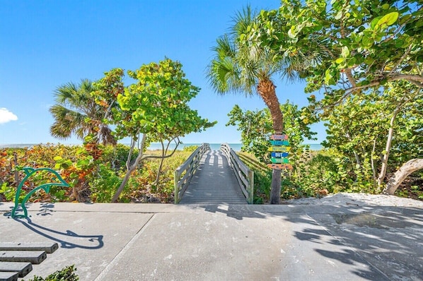 Une promenade en bois mène à une plage bordée de palmiers, et un panneau indique le chemin qui y conduit.