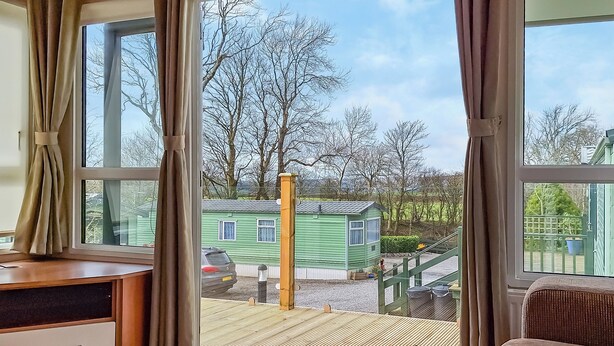 A room with a large window, a wooden desk, a brown sofa, and a view of a green house and trees outside. - 2 bedroom accommodation in Lamplugh (Lamplugh)