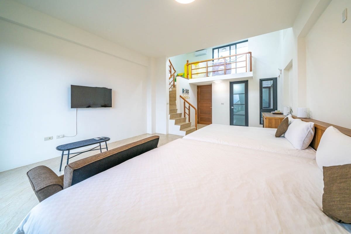 A modern hotel room with a large bed, a desk, a TV mounted on the wall, and a staircase leading to an upper level.