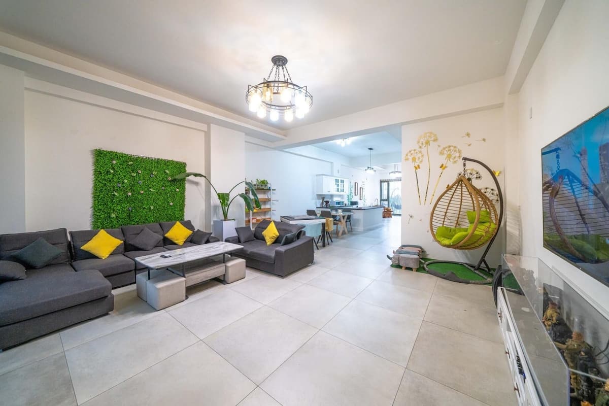 A modern living room with a sectional sofa, a coffee table, and a green wall. There is a kitchen in the background with white cabinets and a hanging chair.