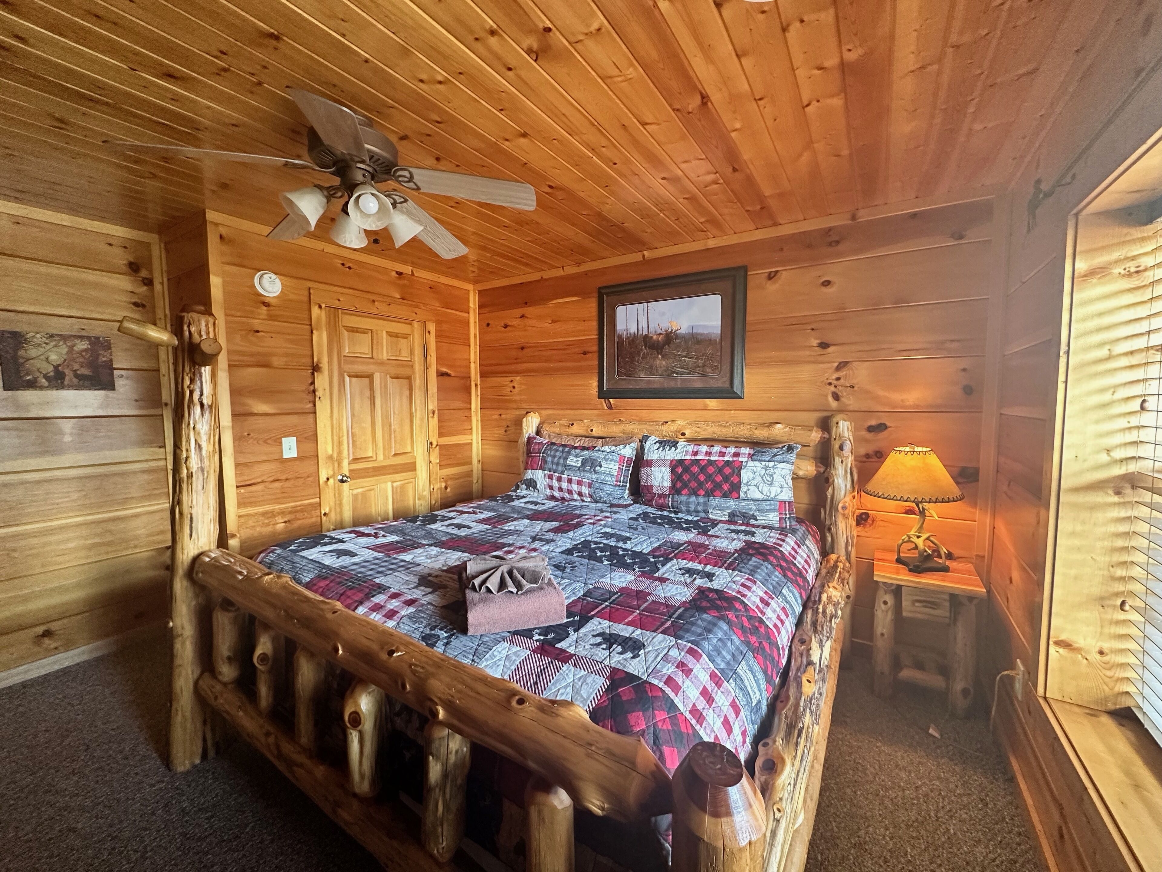 A wooden cabin bedroom with a bed, a ceiling fan, a framed picture, and a nightstand with a lamp.