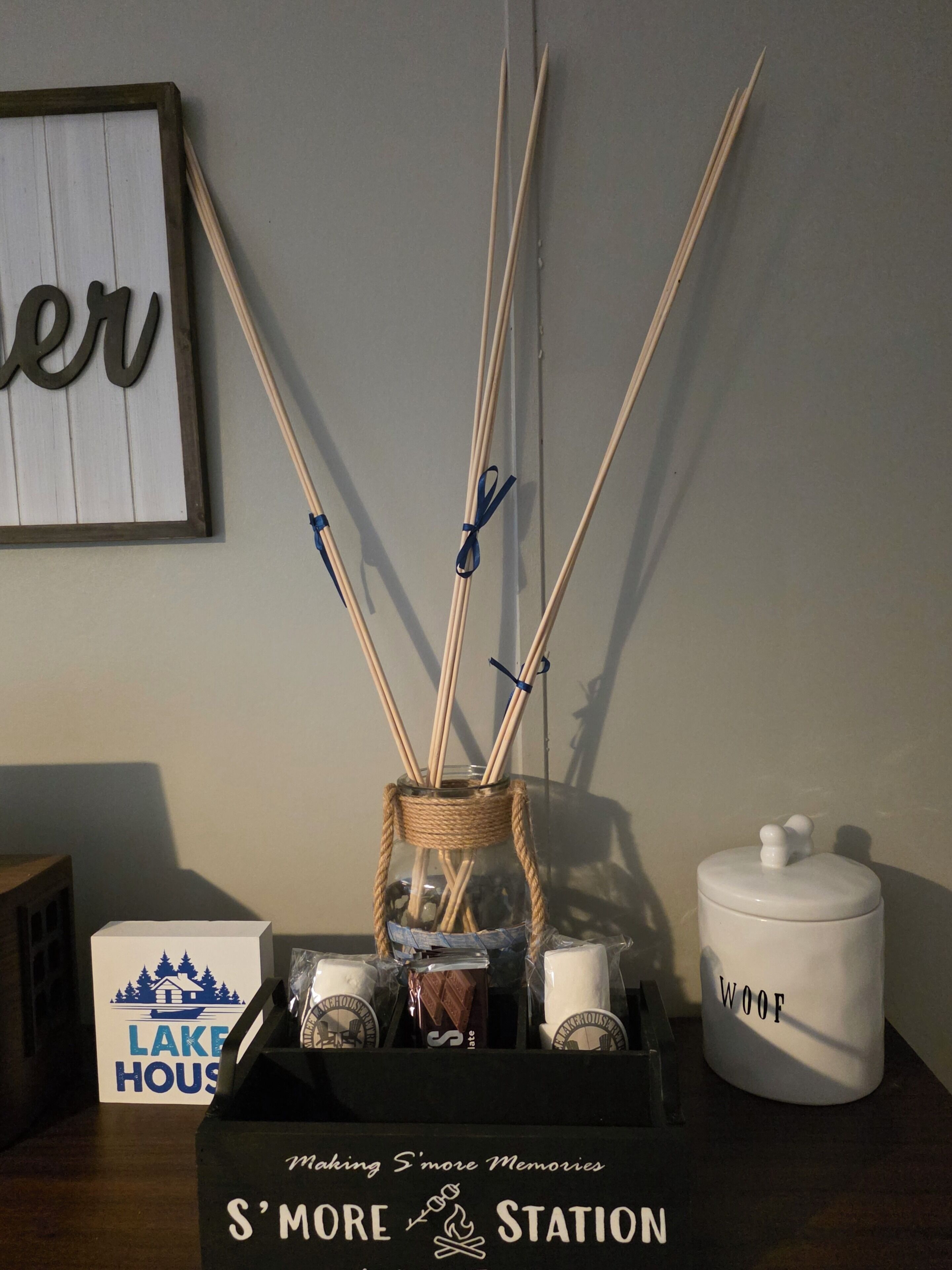 Ein dekorativer Schilferrdiffusor mit rustikaler Vase, einem weißen Behälter mit dem Aufdruck 'WOOF' und einem Schild mit der Aufschrift 'S'MORE STATION'.
