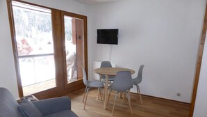 A living room with a grey sofa, a round wooden table with four grey chairs, a flat-screen TV mounted on the wall, and a sliding glass door leading to an outdoor area. - 200m from ski slopes, 3rd floor, balcony, parking, tv, ski locker, 23m², Les Contamines-Montjoie (Les Contamines-Montjoie)