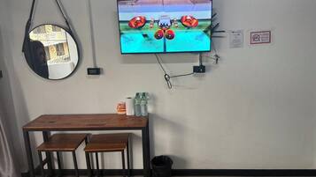 Un televisor montado en la pared mostrando un partido, una mesa de madera con sillas, un cesto de basura y un espejo colgado.