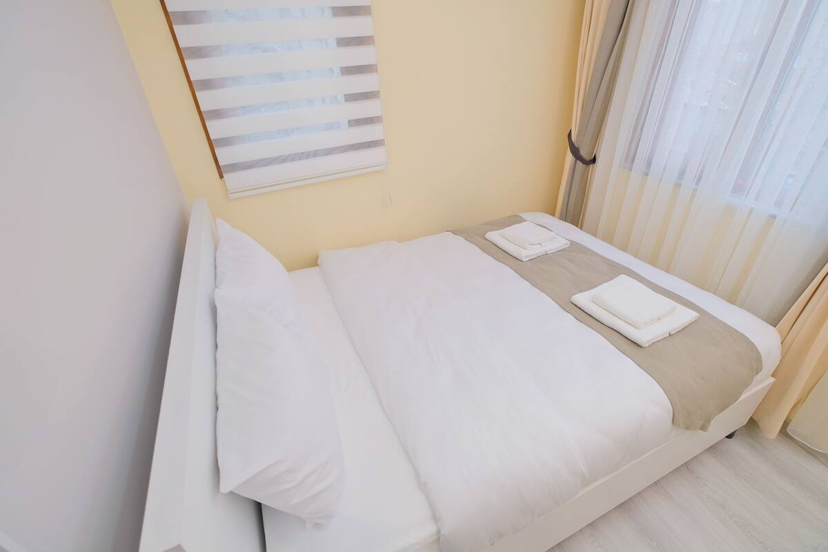 A neatly made bed with white linens and a beige throw blanket, a window with sheer curtains, and a wall-mounted artwork.