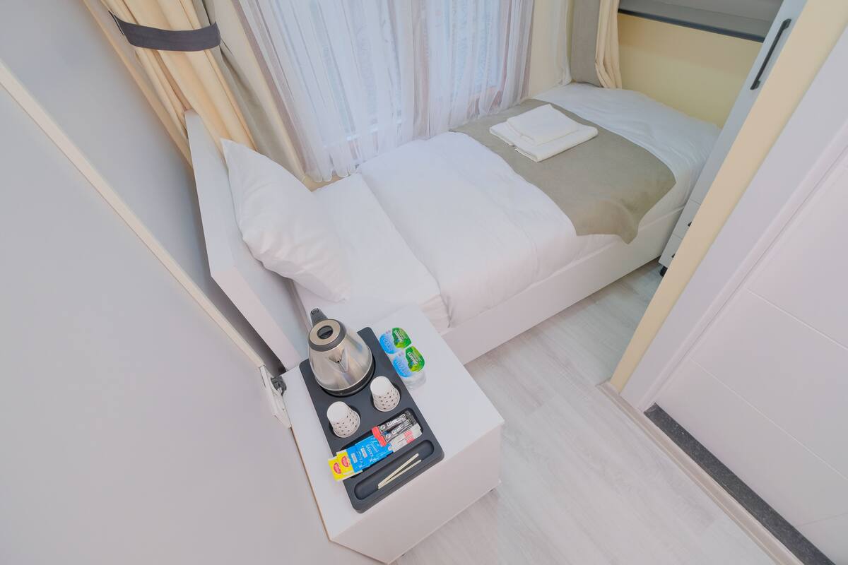 A neatly made bed with a white comforter and pillows, a bedside table with a kettle, tea bags, and a small box of tissues.