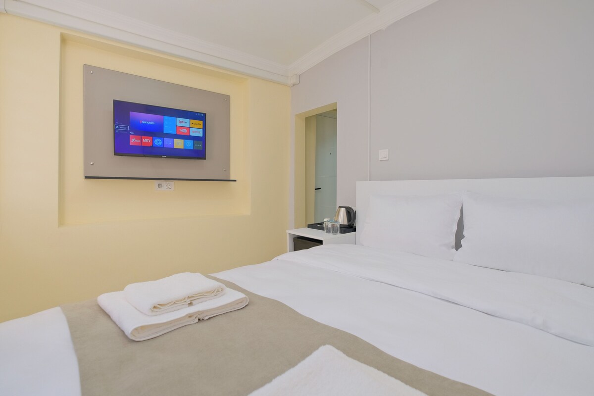 A hotel room with a bed, a TV mounted on the wall, a small table with a kettle and glasses, and a door leading to another room.