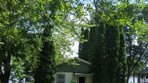 A small, single-story house with a front door and a small porch, surrounded by lush green trees and grass. - Classic Green Lake cottage, lakefront, screened boathouse deck, dock, swimming (Markesan)