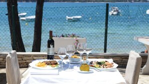 Una mesa de comedor con platos de comida, copas de vino y una botella de vino, con vistas panorámicas al mar y barcos.