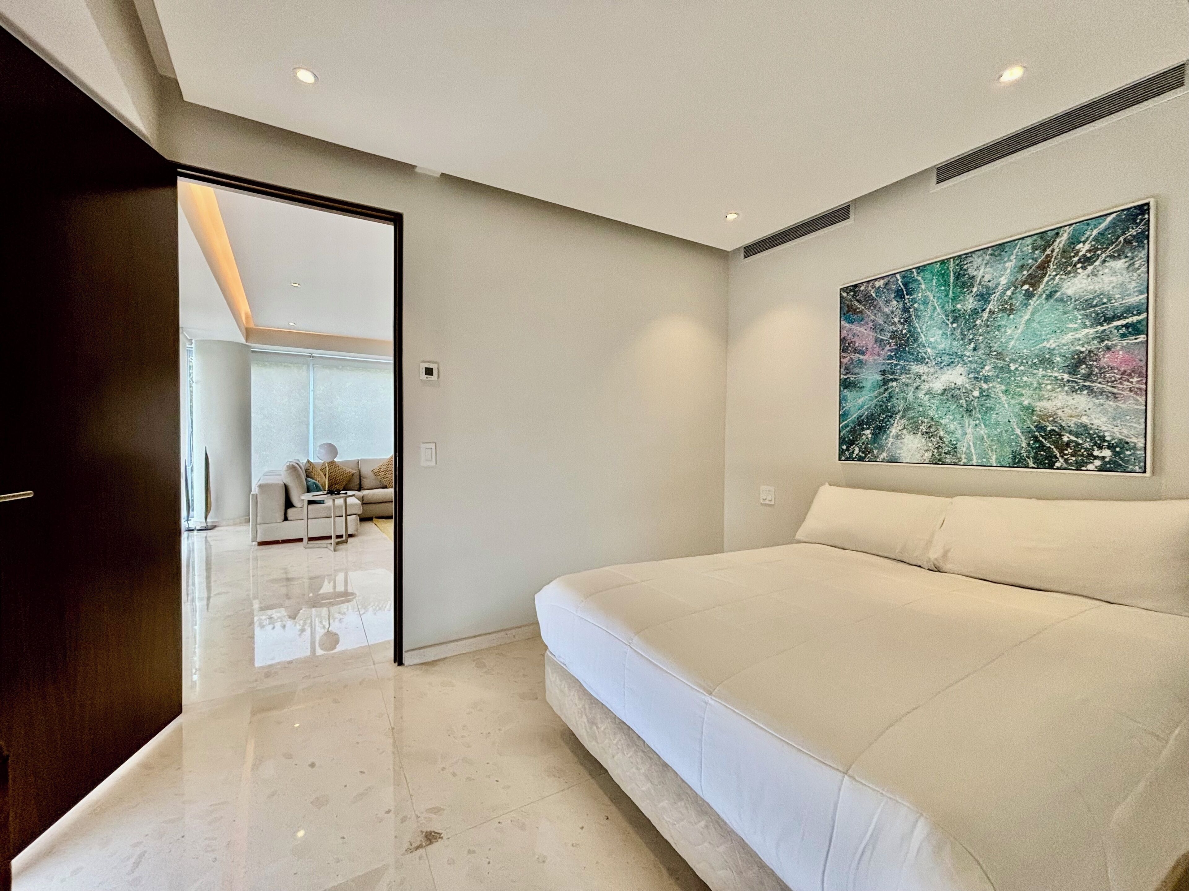 A modern bedroom with a large bed, a painting on the wall, and a view into the living area.