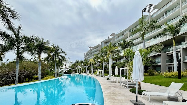 A swimming pool with lounge chairs and umbrellas, surrounded by palm trees and a multi-story building. - Park Life Bolongo (Bucerías)