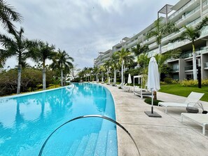 A swimming pool with lounge chairs and umbrellas, surrounded by palm trees and a multi-story building. - Park Life Bolongo (Bucerías)