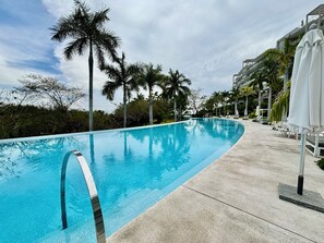 Una piscina de borde curvo, rodeada de palmeras y sillas de descanso, con un edificio al fondo.