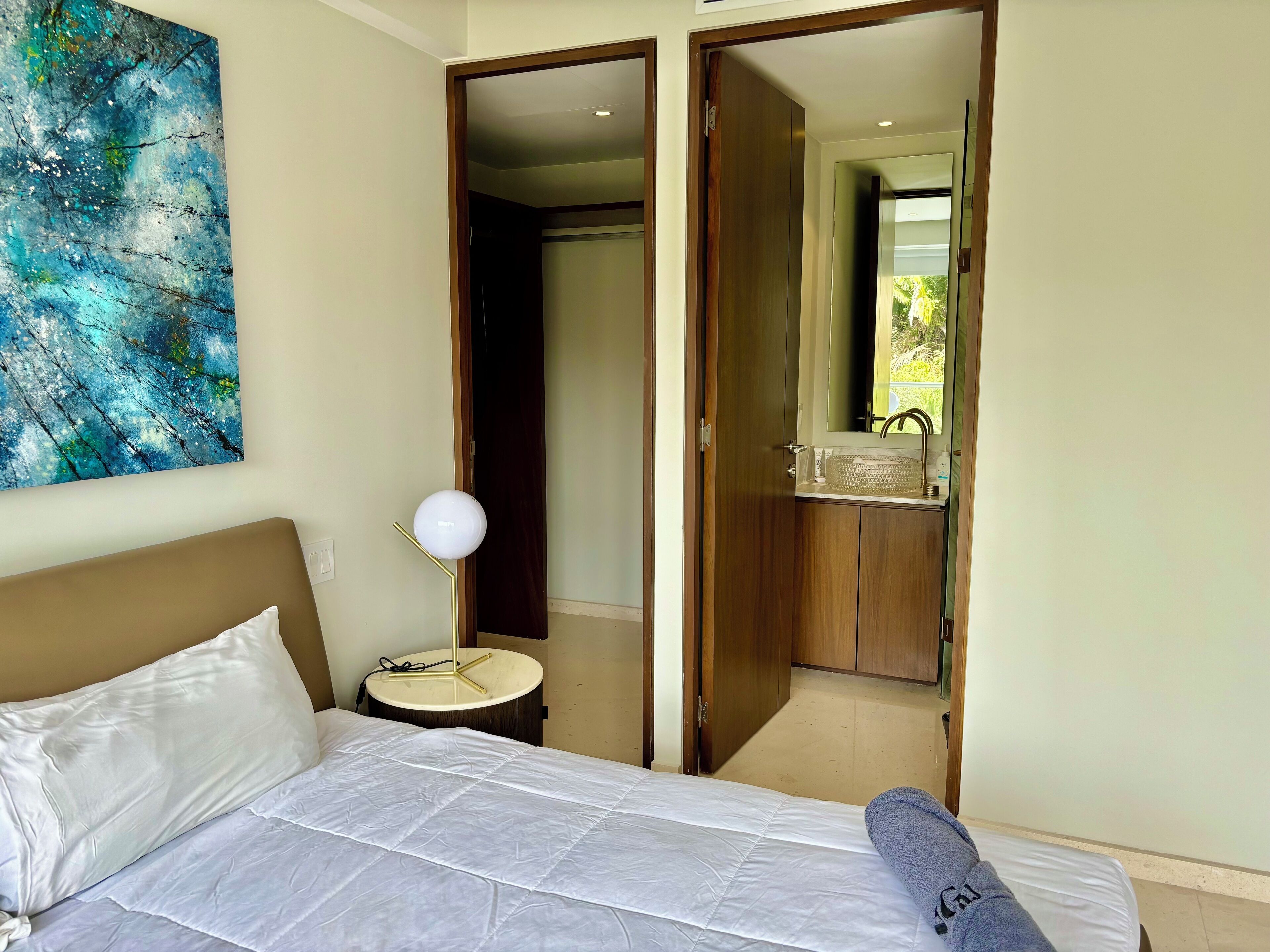 A hotel room with a bed, a bedside lamp, a painting, and an open door leading to a bathroom.