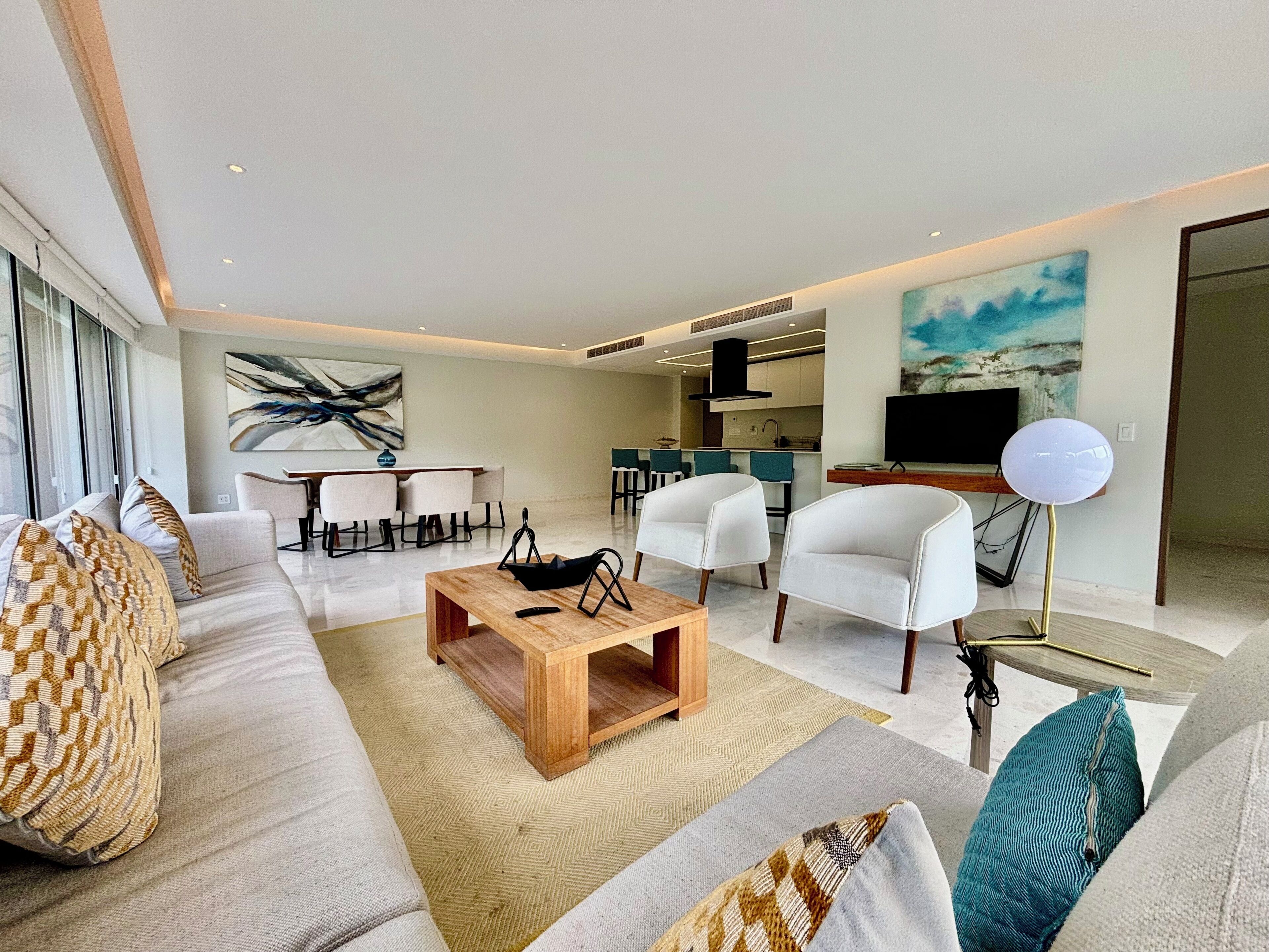 Ein modernes Wohnzimmer mit einer Couch, einem Couchtisch und Sesseln, ein Essbereich mit Tisch und Stühlen sowie eine Küche im Hintergrund.