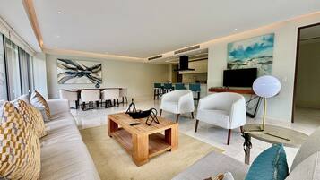 Ein modernes Wohnzimmer mit einer Couch, einem Couchtisch und Sesseln, ein Essbereich mit Tisch und Stühlen sowie eine Küche im Hintergrund.