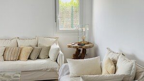 A living room with a white sofa and armchair, a small wooden side table with a lamp, and a window with a view of greenery. - Modern Villa 6 pax, private pool, countryside views, free wifi (Binibeca)