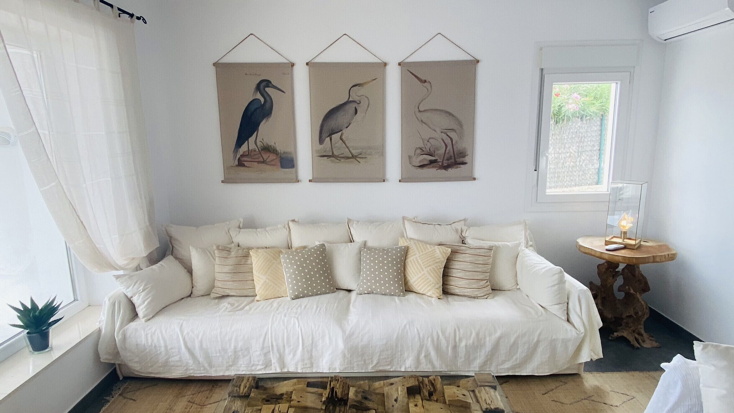 Un sofá blanco con varios cojines, una mesita redonda con una lámpara y tres cuadros de pájaros colgados en la pared.