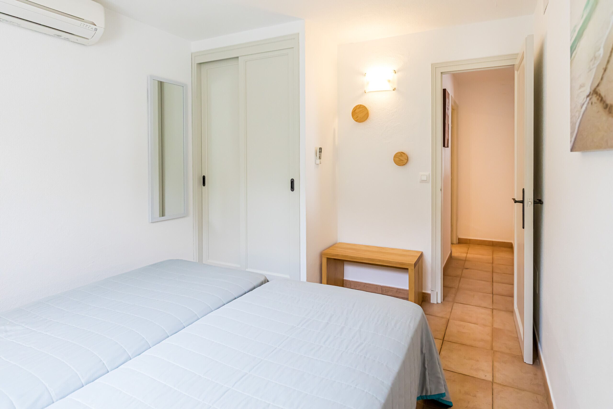 A bedroom with a bed, a bedside table, a wall-mounted light, and a door leading to another room.