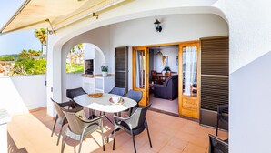 Une terrasse extérieure avec une table ronde et des chaises en osier, un barbecue intégré et une vue sur le jardin.