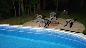 A swimming pool with a tiled edge, surrounded by a grassy area with trees. There are four chairs and a table set up for outdoor dining.