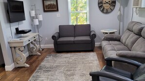 A living room with a grey sofa, a black leather chair, a wooden coffee table, a floor lamp, a TV, and a clock on the wall.