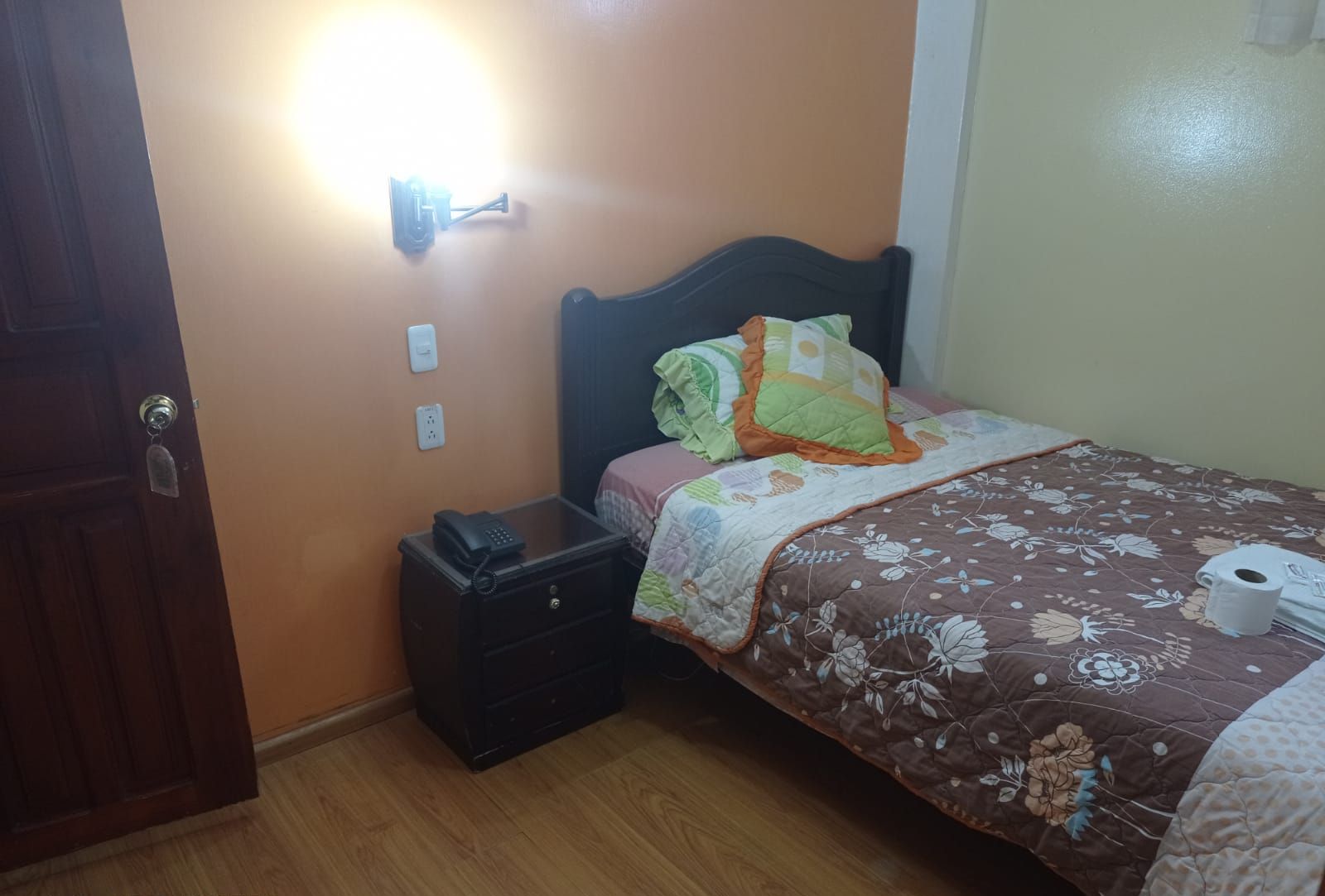 A bedroom with a bed, a nightstand, a wall socket, and a door.